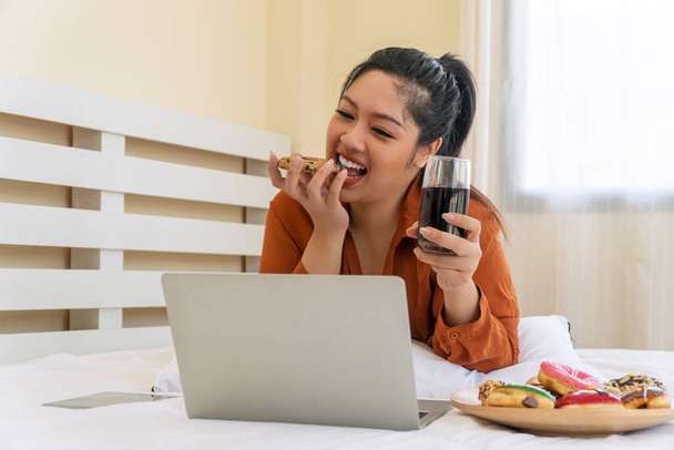 Unhealthy eating and obesity concept. Overweight fat woman holding glass sparkling water with plate doughnut while lying on the with laptop working at home. Fatty girl from sweet donut and soda water. - Photo, Image
