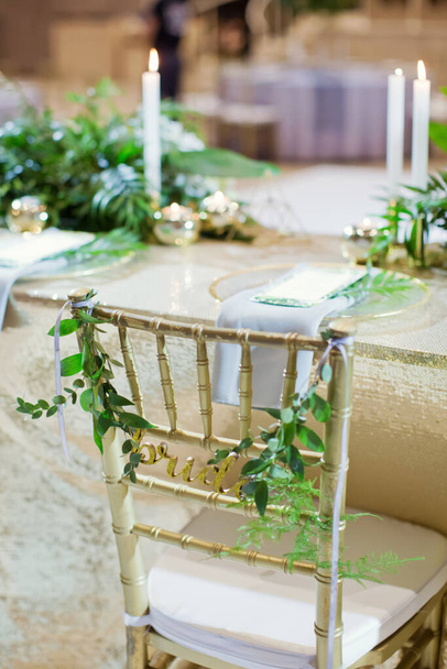 Arany székek táblával menyasszony és vőlegény zöld ruszki gallyakkal. Rendezvényterem arany díszítéssel és trópusi pálmalevéllel - Fotó, kép