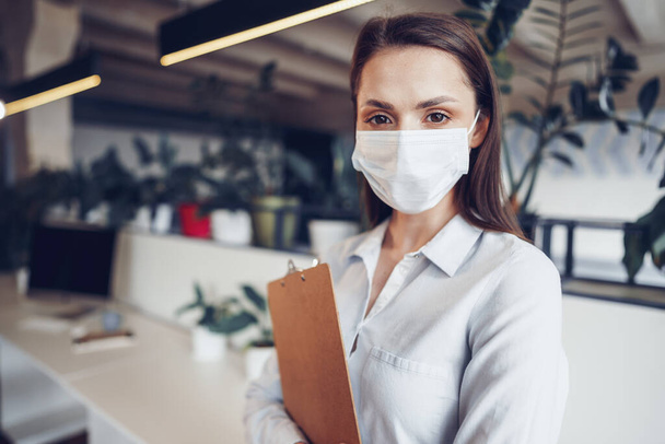 Young businesswoman in face mask standing in office and holding clipboard with documents - Valokuva, kuva