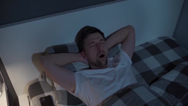Young man looks at the ceiling at night while lying in bed instead of sleeping, checking the time on his mobile phone, suffering from insomnia. Displeased male lying in bed unable to fall asleep - Footage, Video