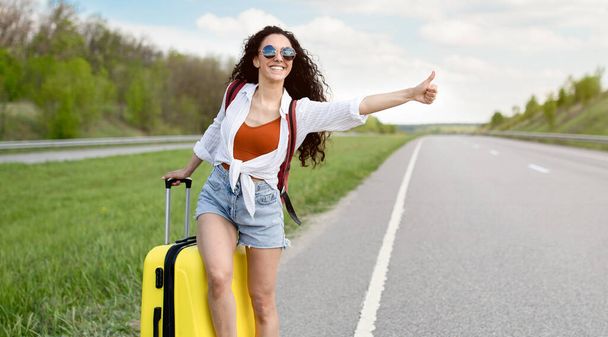 Young lady thumbing her ride, standing with suitcase on highway, trying to catch passing car, hitchhiking in countryside - Photo, Image