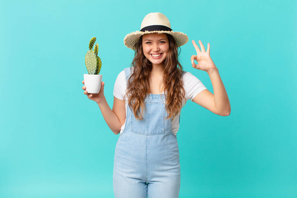 young pretty woman feeling happy, showing approval with okay gesture and holding a cactus - Foto, Bild