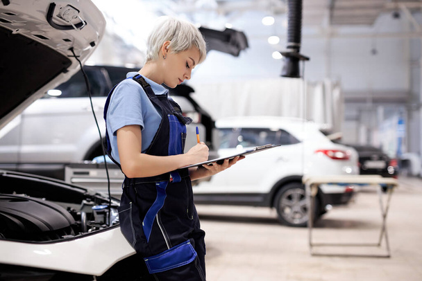 Young technician female concentrated to check list for maintenance in car service - Φωτογραφία, εικόνα