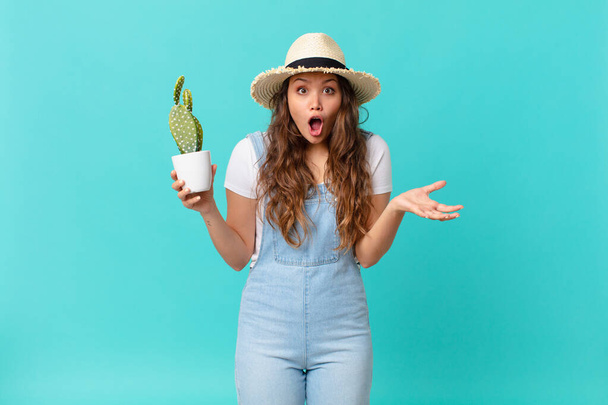 young pretty woman amazed, shocked and astonished with an unbelievable surprise and holding a cactus - Foto, afbeelding