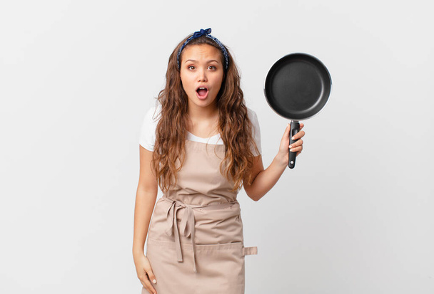 jeune jolie femme regardant concept de chef très choqué ou surpris et tenant une casserole - Photo, image