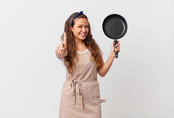 giovane bella donna sorridente orgogliosamente e con fiducia rendendo il concetto di chef numero uno e tenendo una padella - Foto, immagini