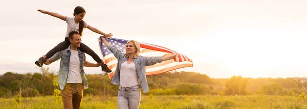 日没のフィールドにアメリカの旗を持つ美しい家族。独立記念日7月4日. - 写真・画像