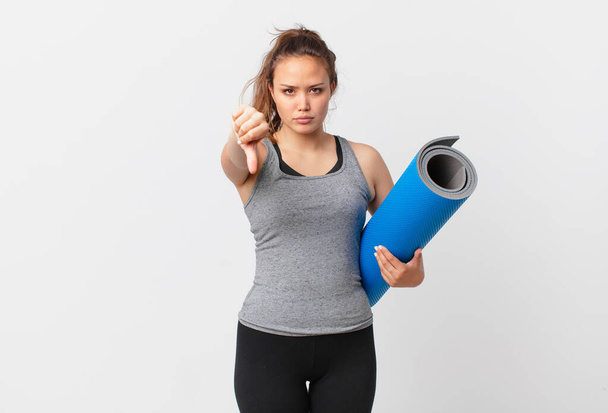 young pretty woman feeling cross,showing thumbs down and holding a yoga mat - Photo, image