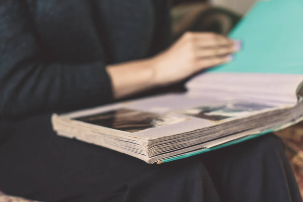 Close-up of a girl in dark clothes sitting on the sofa, holding an album on her lap and looking at old photos - Foto, imagen