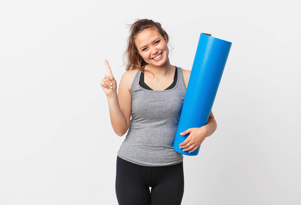 young pretty woman smiling and looking friendly, showing number one and holding a yoga mat - Фото, изображение