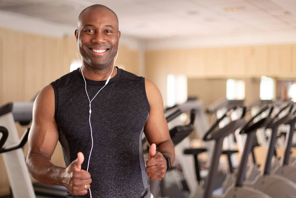 Πορτρέτο του χαμογελαστού μαύρου στο γυμναστήριο. Έχει τους αντίχειρες σε θετική στάση. Έννοια του αθλητισμού και της υγιούς ζωής. - Φωτογραφία, εικόνα
