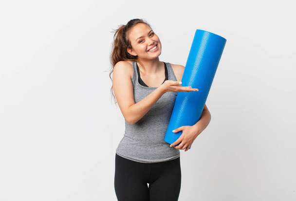 young pretty woman smiling cheerfully, feeling happy and showing a concept and holding a yoga mat - Foto, afbeelding