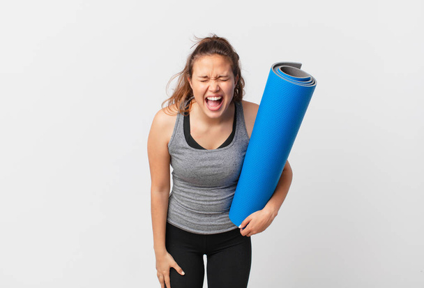 young pretty woman shouting aggressively, looking very angry and holding a yoga mat - Фото, изображение