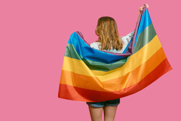 joven desconocida chica rubia caucásica con la espalda vuelta, sosteniendo una bandera de arco iris sobre un fondo rosado. concepto de arco iris, símbolo de LGBTQIA gay-lesbiana orgullo y diversidad. - Foto, Imagen