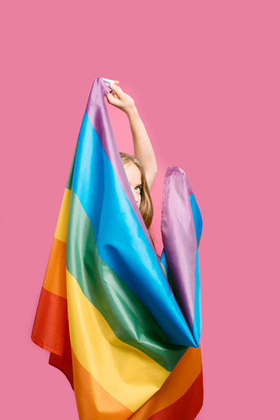 young blonde Caucasian girl covering her face with a rainbow flag on a pink background.Rainbow concept, symbol of LGBTQIA gay-lesbian pride and diversity. vertical image. copy space. - Foto, afbeelding