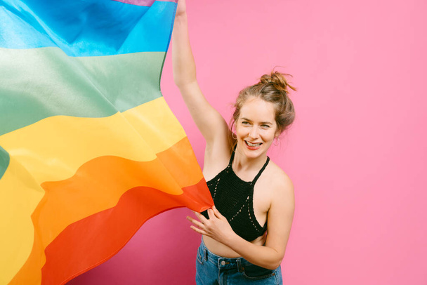 young blonde Caucasian girl playing with a rainbow flag on a pink background.Rainbow concept, symbol of LGBTQIA gay-lesbian pride and diversity. - 写真・画像