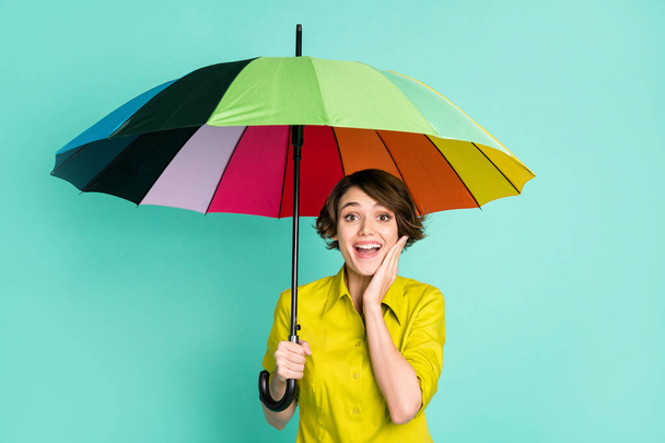 Photo portrait of amazed smiling girl touching cheek keeping parasol isolated on vibrant teal color background - Photo, Image