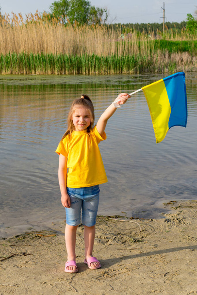 Ukrayna bayrağı Ukraynalı küçük bir kızın ellerinde bir gölün ve sazlıkların arkasında dalgalanıyor. Ukrayna ve Bayrak Günü 'nün Bağımsızlık Günü. - Fotoğraf, Görsel