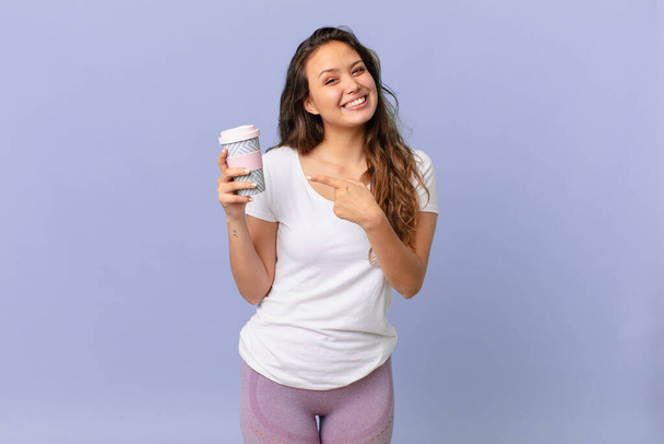 young pretty woman smiling cheerfully, feeling happy and pointing to the side and holding a coffee - Foto, Bild