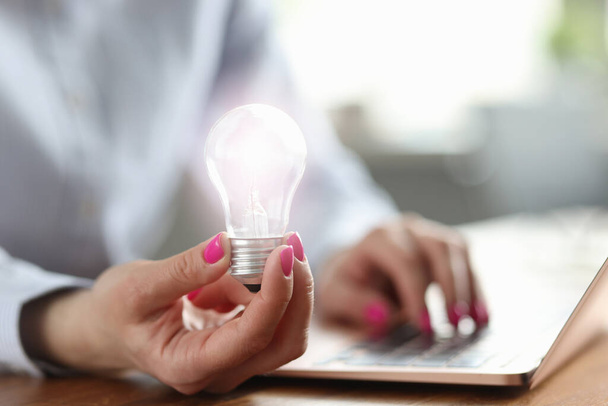 Femme tient ampoule dans sa main et travaille sur ordinateur portable - Photo, image