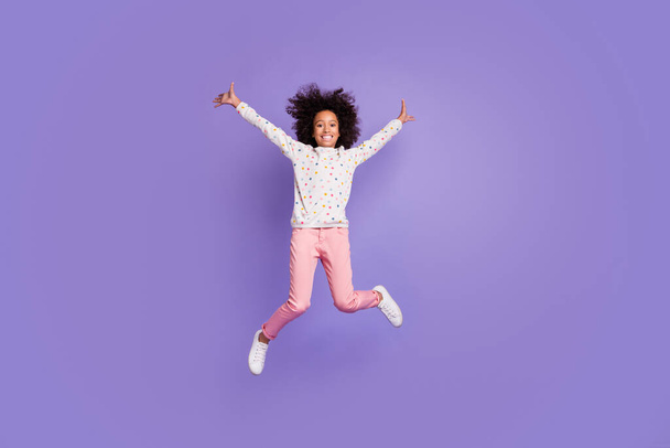 Full size photo of happy afro american little girl jump wear pink pants white jumper isolated on purple color background - Zdjęcie, obraz