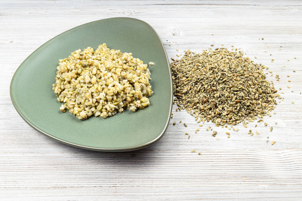 Сырые зерна пшеницы и вареная каша на зеленой тарелке на деревянном столе - Фото, изображение