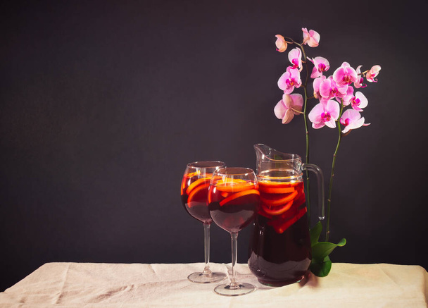 Czerwone wino sangria z owocami w szklankach i dzbanek na stole. Fioletowa orchidea blisko. Mroczne tło. Widok z miejsca kopiowania. - Zdjęcie, obraz