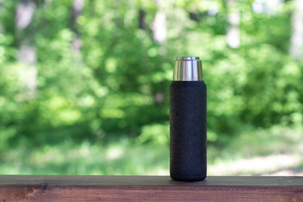 A black thermos with brewed tea or coffee stands on a wooden board with a copyspace. Thermos with hot drink - Fotó, kép