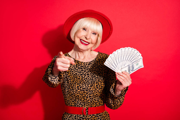 Photo of aged woman happy positive smile hold cash dollars rich indicate finger you isolated over red color background - Foto, Imagem