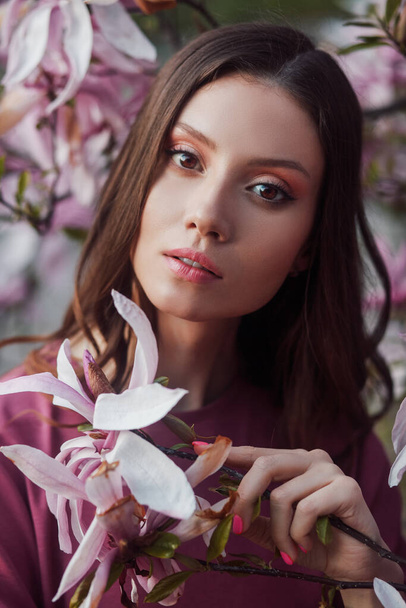 マグノリアの花の背景に立つ美人。庭に春の花を持つ若い女性。本物の肖像画だ。肖像画だ。魅力的な女性のクローズアップ肖像画  - 写真・画像