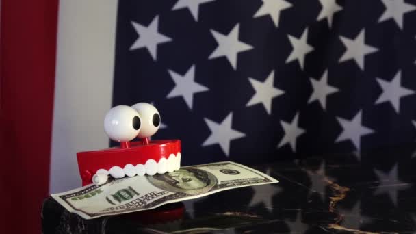 Una prótesis de juguete sostiene un billete de dólar en los dientes contra el fondo de la bandera estadounidense . - Metraje, vídeo