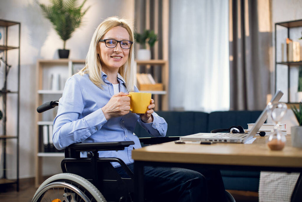 Woman in wheelchair holding cup of coffee and using laptop - Zdjęcie, obraz
