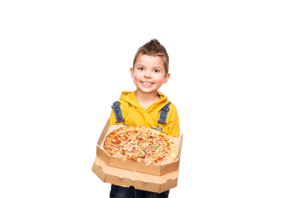 Malý roztomilý chlapeček ve žlutém svetru a džínové kombinézy se usmívá a drží otevřenou krabici s pizzou izolované na pozadí - Fotografie, Obrázek