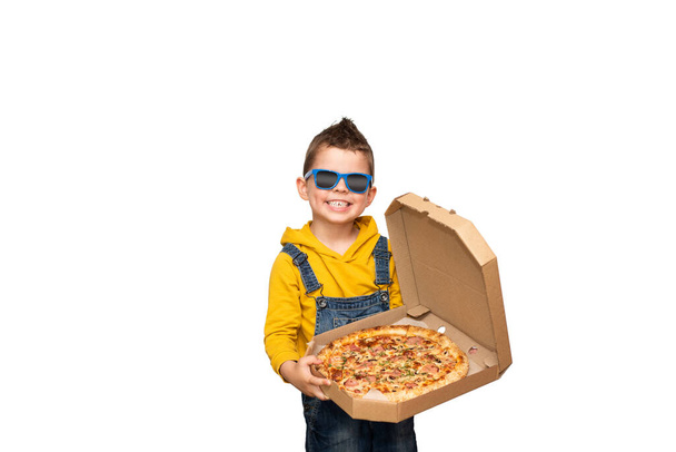 Kleine schattige baby jongen in een gele trui en denim overall en blauwe zonnebril glimlacht en houdt een open doos met pizza geïsoleerd op de achtergrond. - Foto, afbeelding