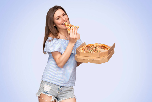 Linda jovem mulher sorridente em um chapéu de palha e top azul com um decote comendo uma fatia de pizza e segurando uma caixa aberta com uma grande pizza isolada no fundo. - Foto, Imagem