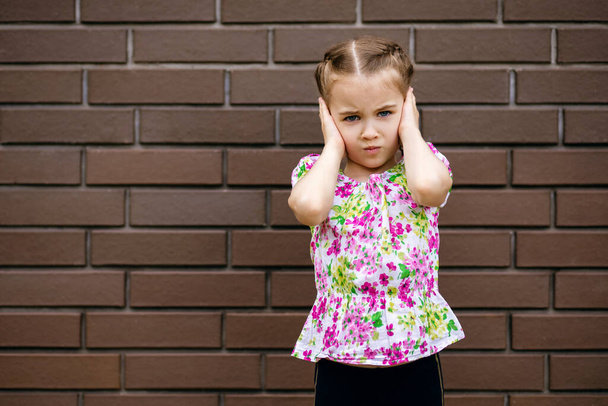 Mała dziewczynka emocjonalnie zamyka uszy stojąc przy ceglanym murze. Słodkie dziecko nie lubi głośnych hałasów i wścieka się. Wolne miejsce na tekst. Jasne letnie ubrania niebieskookiej dziewczyny - Zdjęcie, obraz