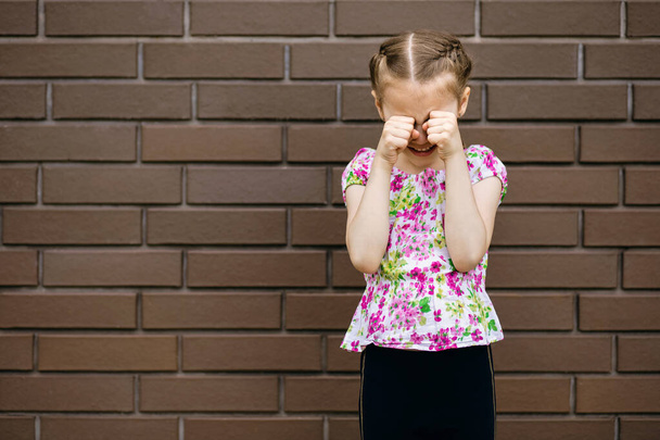 A little girl with a beautiful hairstyle is very upset and cries, wiping her eyes with her hands. Preschool child is very upset and crying against the background of a brick wall with a place for text - Photo, Image