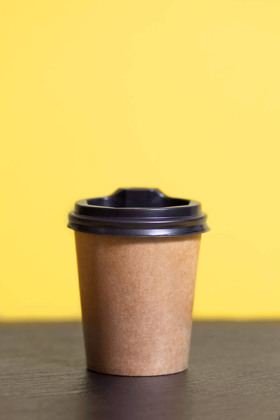 Одноразовая кофейная чашка для кафе на желтом фоне. Коричневый картонный макет экологически чистой чашки кофе на вертикальной фотографии. Одноразовые пластмассовые и бумажные горячие напитки шаблон - Фото, изображение