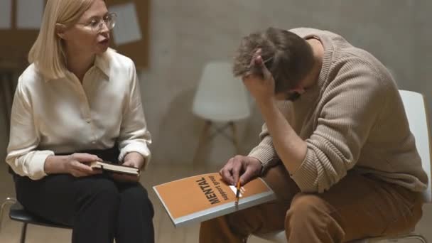 Medium laukaus naispsykologi tukee emotionaalinen mies masennus koskettaa hiuksiaan ottaa istunnon kuntoutuskeskuksessa - Materiaali, video