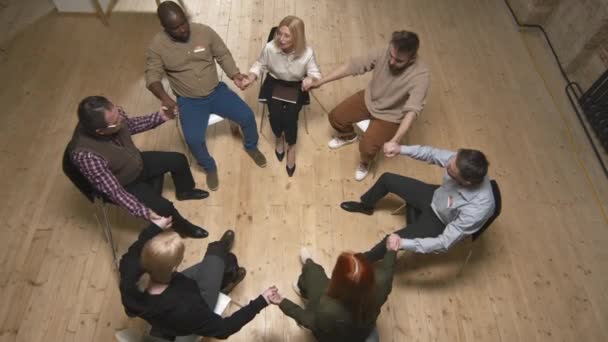 Top-view shot of small group of people having mental problems and female psychologist sitting in circle and holding hands together supporting each other - Footage, Video