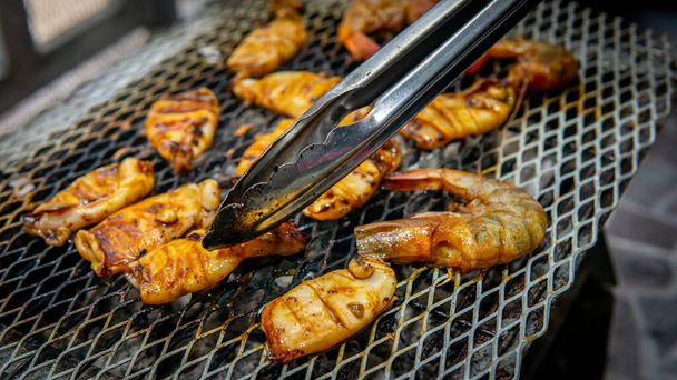 Close up view of marinated cutlet squids and shrimps cook and flip using a stainless steel tongs on a hot grill during the barbeque party. Smoky and juicy seafood concept. - Photo, Image