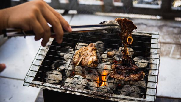 Piknik partisinde sıcak dumanlı kömür ızgarasında pişirilmiş metal maşayla sosla marine edilmiş çiğ kuzu eti tutan eli yakından izleyin. Önplanda seçmeli odak. - Fotoğraf, Görsel