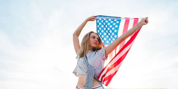 Νεαρή ξανθιά γυναίκα της χιλιετίας στέκεται με αμερικανική σημαία δίπλα στον γαλάζιο ουρανό. Σημαία των Ηνωμένων Πολιτειών στα χέρια της. 4η Ιουλίου Ημέρα Ανεξαρτησίας. Εθνική εορτή των ΗΠΑ. Έννοια ελευθερίας και μνήμης - Φωτογραφία, εικόνα
