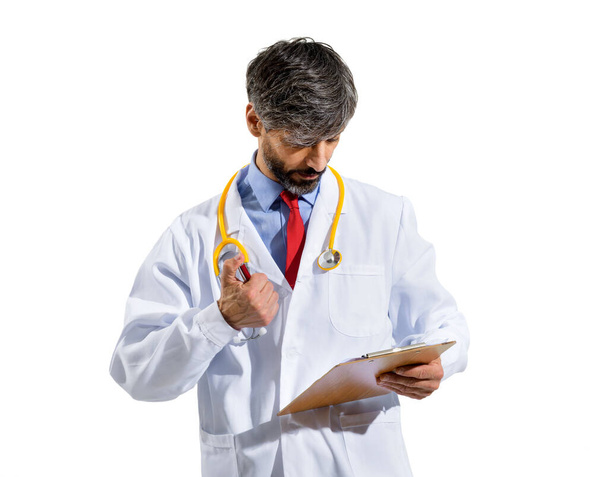 Arzt in weißem Laborkittel und Stethoskop, der Patientenakten oder Notizen auf einem Klemmbrett mit ernstem Gesichtsausdruck vor weißem Studiohintergrund aufbewahrt - Foto, Bild