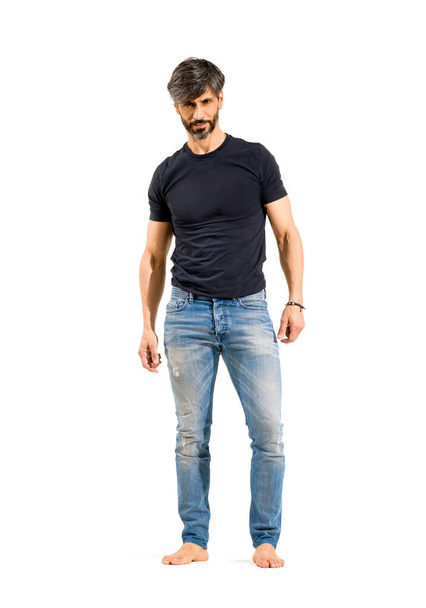 50-jarige pasvorm trendy casual man staande kijken naar camera in jeans en blote voeten met een rustige vriendelijke glimlach geïsoleerd over een witte studio achtergrond - Foto, afbeelding