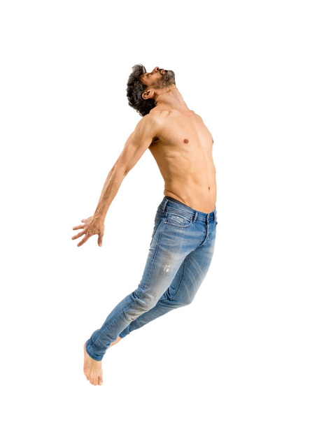 Fit γυμνόστηθη ξυπόλυτος άνθρωπος σε τζιν πηδώντας στον αέρα καμπυλώνοντας το κεφάλι του πίσω με τα χέρια επεκταθεί απομονωμένο πάνω από ένα λευκό φόντο στούντιο - Φωτογραφία, εικόνα