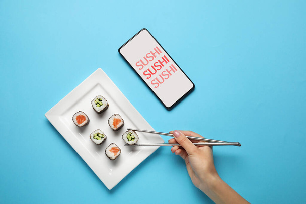 Female hand with chopsticks, sushi rolls on plate and mobile phone on color background - Foto, Imagen
