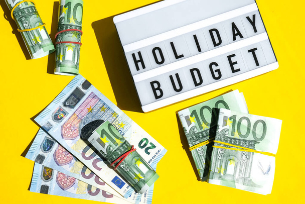 Lightbox board met woord HOLIDAY BUDGET op gele achtergrond rond eurobankbiljetten. Geld, zaken, financiën, investeringen, sparen. Contante rekening. Bedrijfsbegroting van rijkdom en welvaartsfinanciering. Passief inkomen - Foto, afbeelding