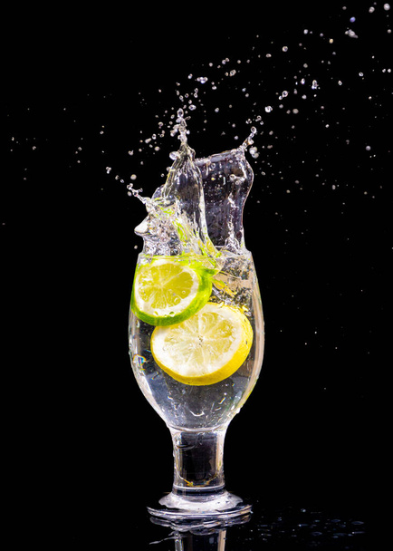 A vertical shot of  lemon slices splashing into glass water on isolated black background - Foto, afbeelding