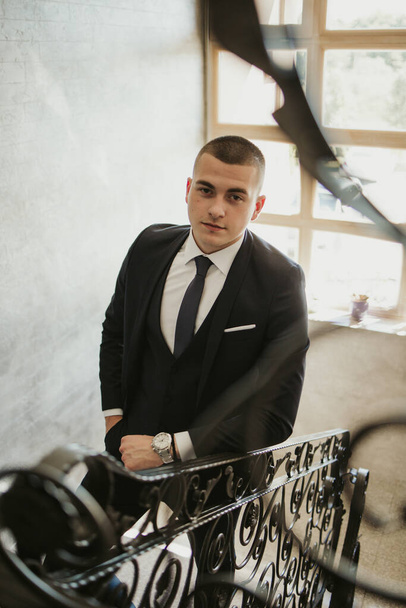 A vertical shot of a young caucasian man in a black suit leaning on the handrail - Foto, Imagen